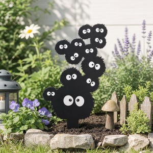 May include: A black garden stake featuring a cluster of multiple fuzzy soot sprite characters, each with two large white eyes and black pupils. The design is vertically oriented with a jagged, circular texture, creating a whimsical stack of creatures.