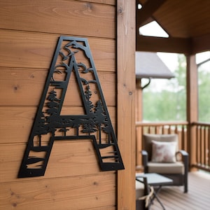 May include: Black metal letter "A" with a nature scene cut-out design. The design includes trees, mountains, birds, and a sun. The letter is mounted on a wooden wall, suitable for home decor.