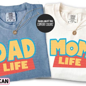 two t-shirts with the text "DAD LIFE" printed on them, one in blue and one in white.