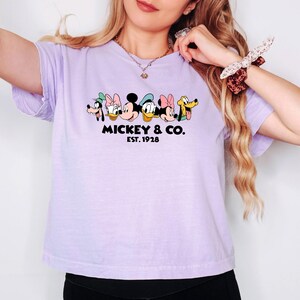 A woman wearing a purple t-shirt with a graphic of Mickey Mouse and Minnie Mouse, along with the text "MICKEY & CO. EST. 1928".