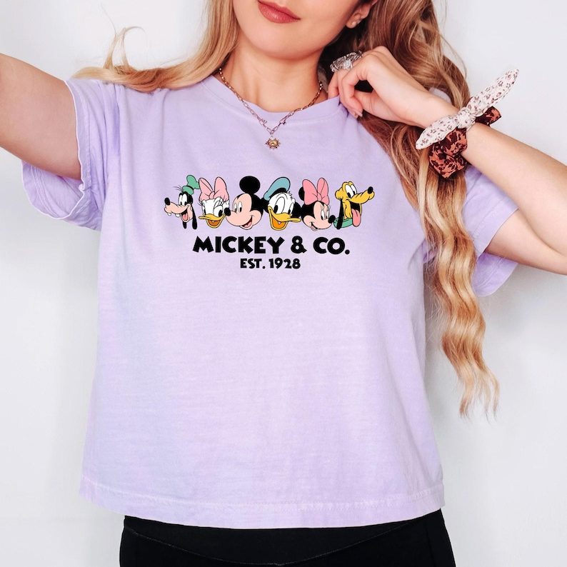 A woman wearing a purple t-shirt with a graphic of Mickey Mouse and Minnie Mouse, along with the text "MICKEY & CO. EST. 1928".