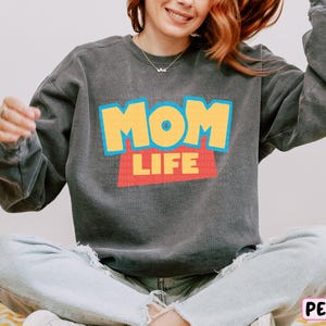 A woman wearing a gray sweatshirt with the text "MOM LIFE" printed on it, sitting cross-legged on the floor with her arms outstretched.