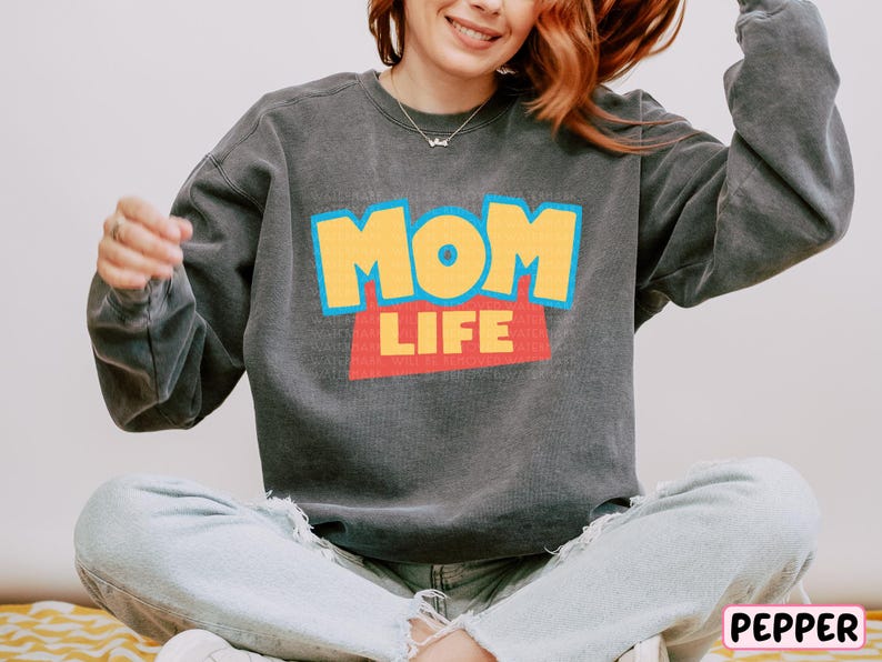 A woman wearing a gray sweatshirt with the text "MOM LIFE" printed on it, sitting cross-legged on the floor with her arms outstretched.