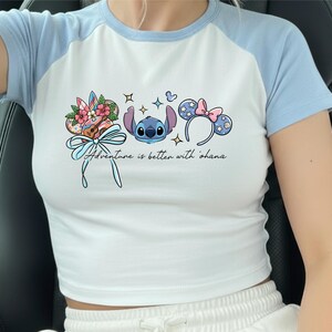 A person wearing a white and blue cropped top with a graphic design featuring a character from the Disney movie Lilo and Stitch, along with various Disney-themed elements such as flowers and earmuffs.