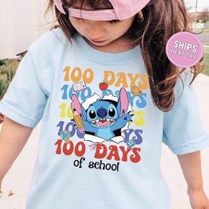A young girl wearing a light blue t-shirt with a colorful graphic design featuring a cartoon character and the text "100 DAYS of school".