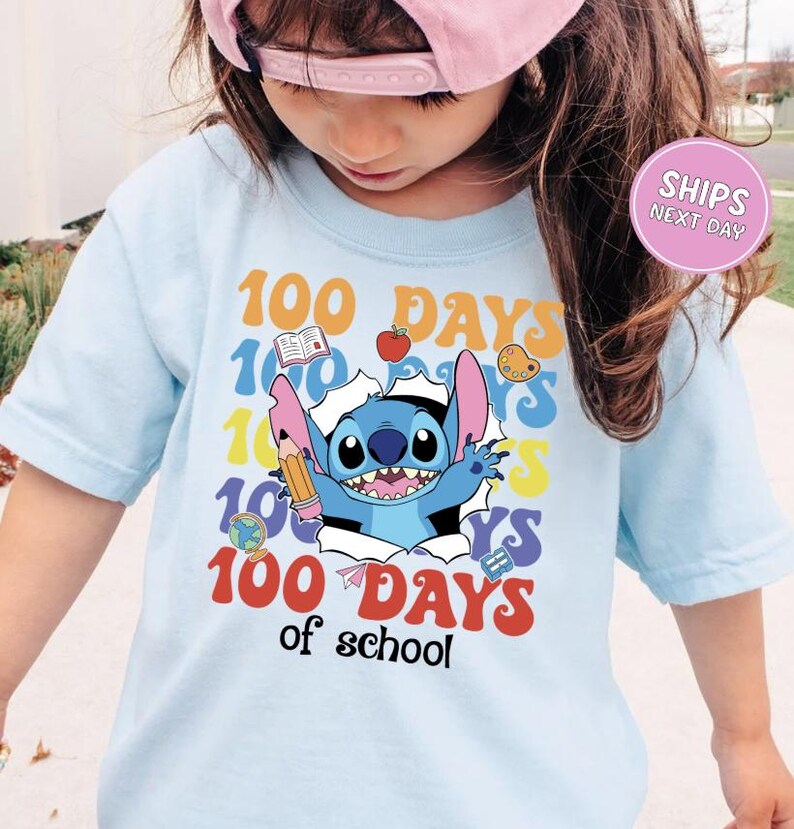 A young girl wearing a light blue t-shirt with a colorful graphic design featuring a cartoon character and the text "100 DAYS of school".