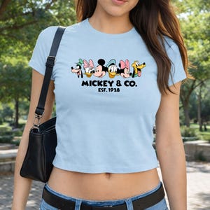 A woman wearing a light blue crop top with a graphic of Mickey Mouse and Donald Duck on it, along with the text "MICKEY & CO. EST. 1978".
