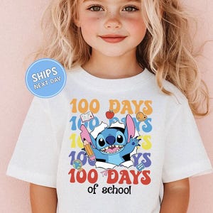 A young girl wearing a white t-shirt with a graphic design featuring a cartoon character and the text "100 DAYS of school".