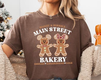 May include: A brown t-shirt featuring a graphic of two gingerbread people with Mickey and Minnie Mouse ears and bows. The text on the shirt reads "Mickey & Minnie's Main Street Bakery Cookies Gingerbread Cake."