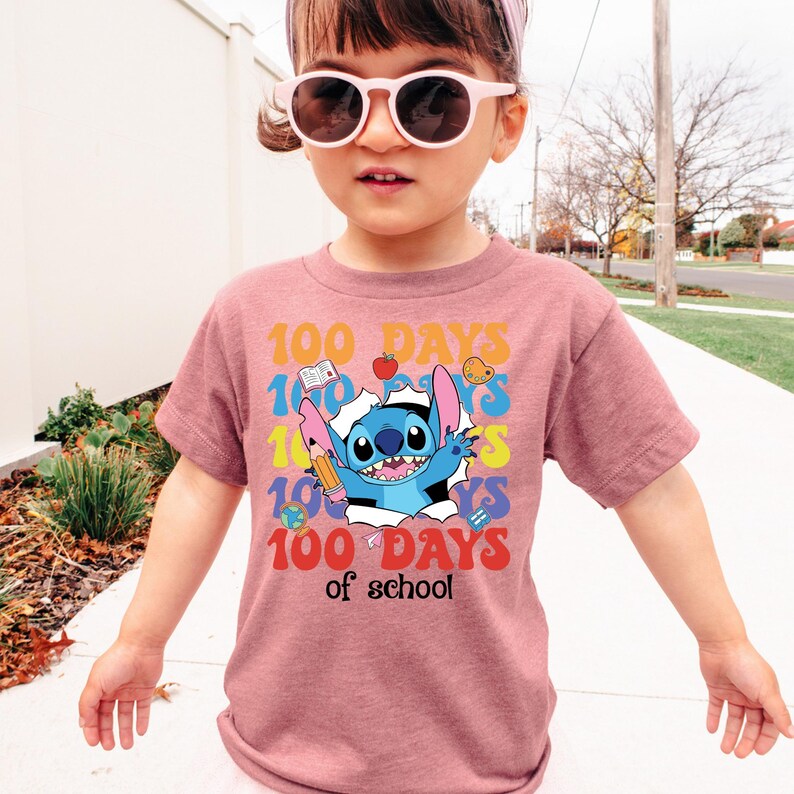 A young girl wearing a pink t-shirt with a cartoon character and the text "100 DAYS of school" printed on it.
