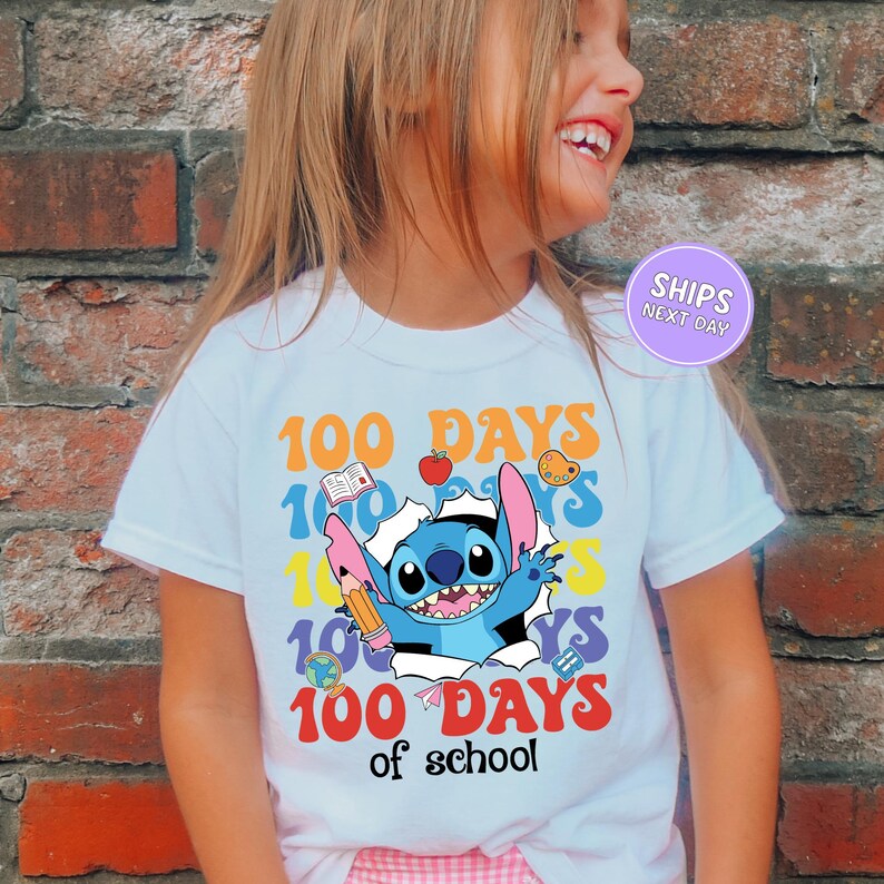 A young girl wearing a white t-shirt with a colorful graphic design featuring a cartoon character and the text "100 DAYS of school".