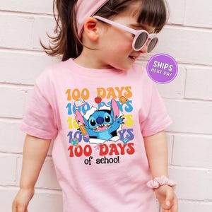 A young girl wearing a pink t-shirt with a graphic design of a cartoon character and text celebrating 100 days of school.