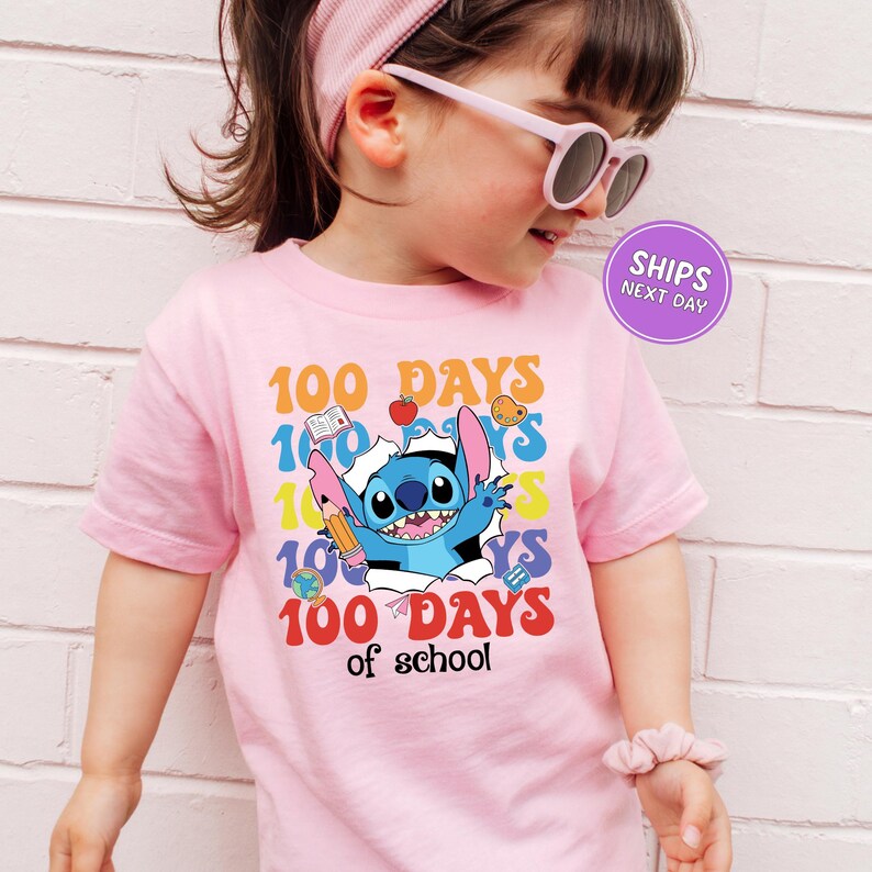 A young girl wearing a pink t-shirt with a graphic design of a cartoon character and text celebrating 100 days of school.