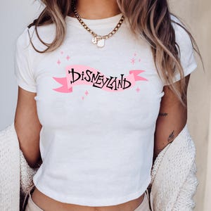 A woman wearing a white t-shirt with the word "Disneyland" printed on it, along with a necklace and a white sweater.