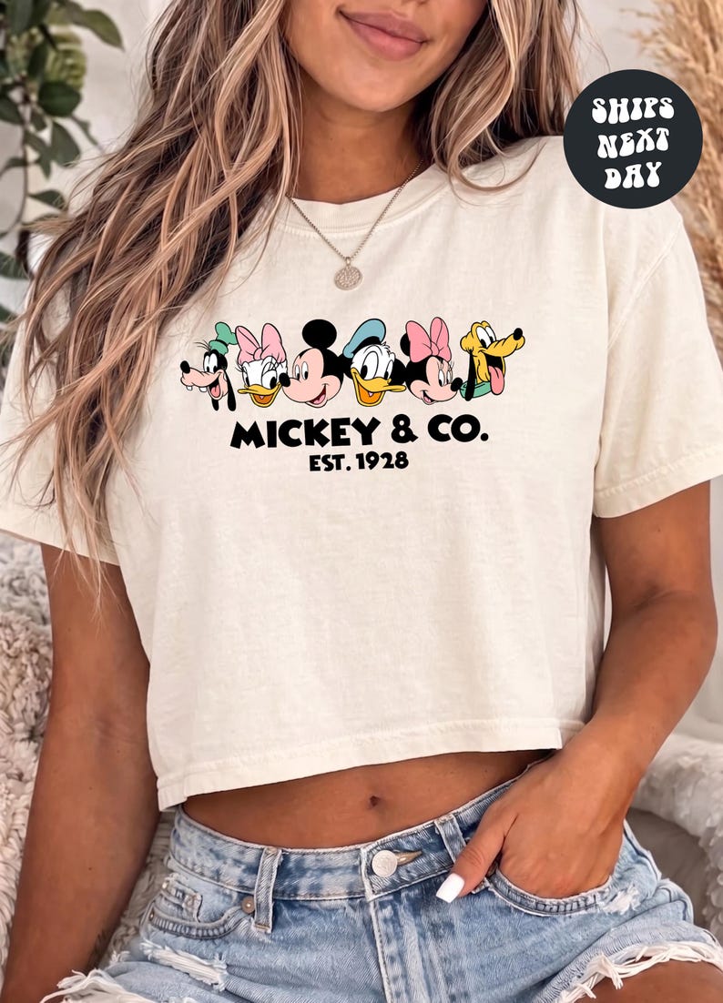 A woman wearing a white crop top with a graphic of Mickey Mouse and Donald Duck, along with the text "Mickey & Co. EST. 1928".