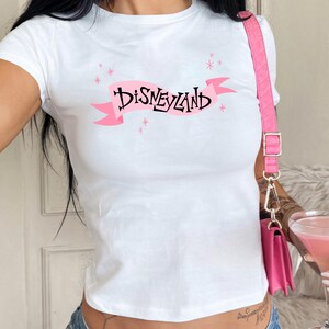A woman wearing a white t-shirt with the word "Disneyland" printed on it, holding a pink drink in her hand.