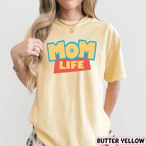 A woman wearing a yellow t-shirt with the text "MOM LIFE" printed on it.