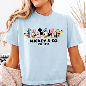 A woman wearing a light blue t-shirt with a graphic design featuring the faces of Disney characters Mickey Mouse, Minnie Mouse, Donald Duck, Goofy, and Pluto.
