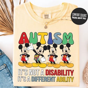 May include: A light yellow Comfort Colors sweatshirt featuring the word "AUTISM" in colorful letters above four Mickey Mouse figures. Below the characters, the text reads "IT'S NOT A DISABILITY IT'S A DIFFERENT ABILITY."