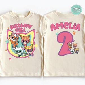 May include: A cream-colored t-shirt featuring a colorful graphic of cartoon cats. The front of the shirt says "BIRTHDAY GIRL" and the back says "AMELIA 2". The design includes paw prints and stars.