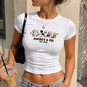 A woman wearing a white t-shirt with a Disney character graphic and text, holding a drink in her hand.
