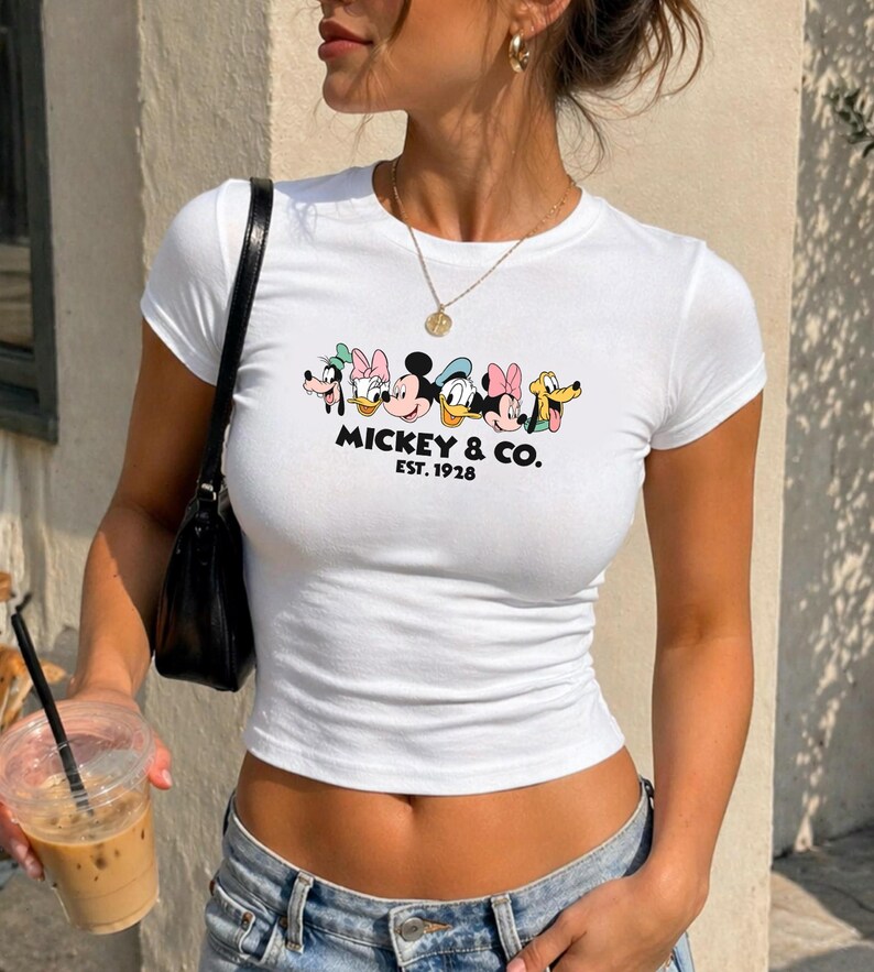 A woman wearing a white t-shirt with a Disney character graphic and text, holding a drink in her hand.