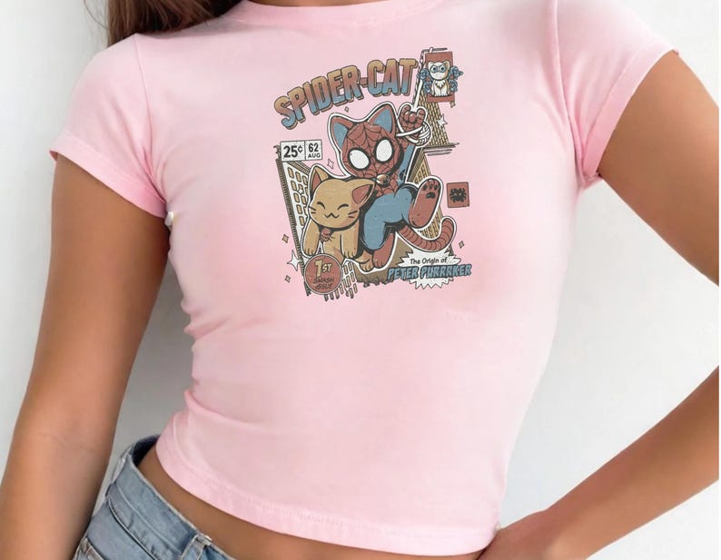 A pink crop top with a graphic design featuring a cat and a spider-like character, along with text and various other elements.