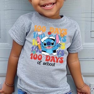 A young child wearing a gray t-shirt with a colorful graphic design featuring a cartoon character and the text "100 DAYS of school".