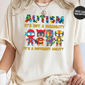 May include: Cream-colored t-shirt with the word "AUTISM" in colorful puzzle piece letters. Below, the text reads "IT'S NOT A DISABILITY, IT'S A DIFFERENT ABILITY." Four cartoon superheroes are featured below the text.
