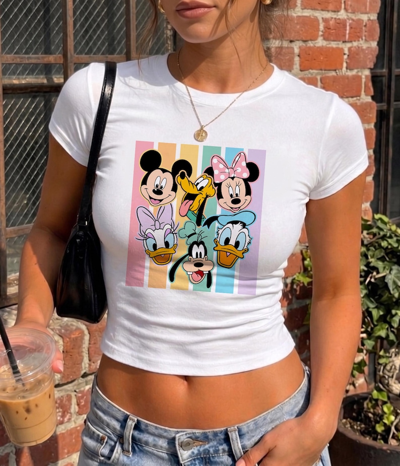 A woman wearing a white t-shirt with a graphic of Disney characters Mickey Mouse, Minnie Mouse, Donald Duck, Goofy, and Pluto. She is holding a drink in her hand.
