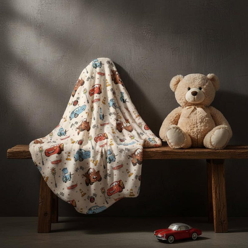 May include: A cream-colored baby blanket with a repeating pattern of cartoon cars, traffic lights, and road signs. A beige teddy bear sits next to the blanket on a wooden bench. A small red toy car is in the foreground.