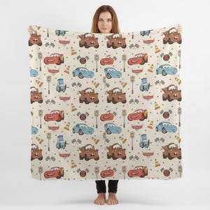 May include: A beige blanket featuring a repeating pattern of cartoon cars, traffic lights, and checkered flags. The cars are in red, blue, and brown. The blanket is held up by a person.