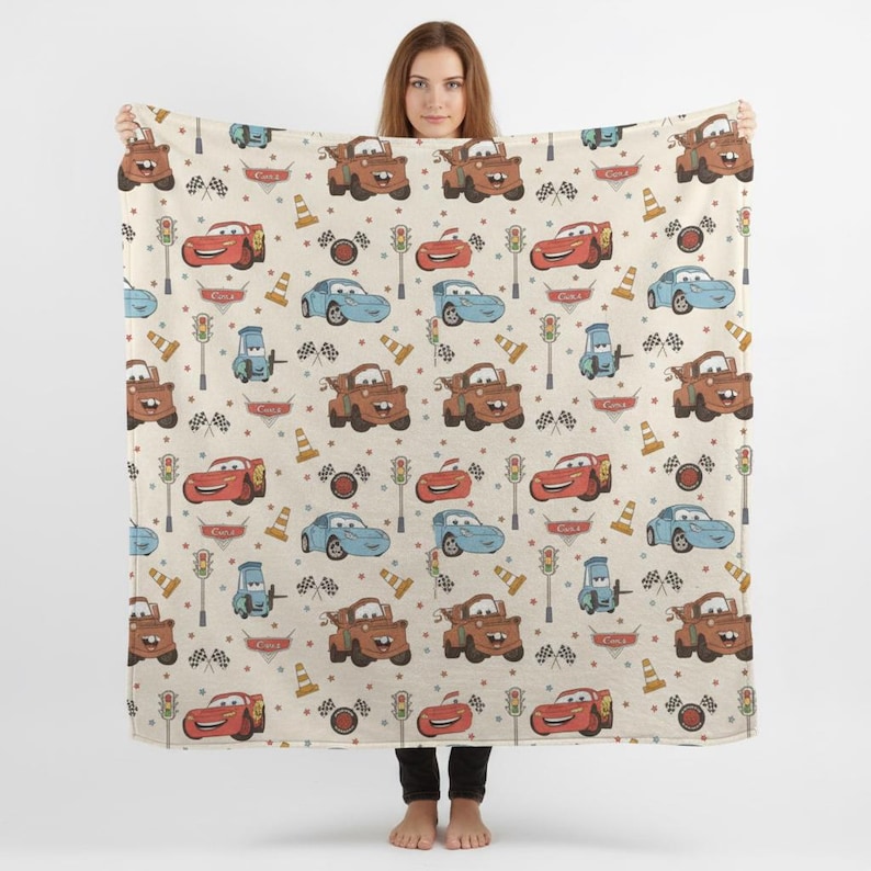 May include: A beige blanket featuring a repeating pattern of cartoon cars, traffic lights, and checkered flags. The cars are in red, blue, and brown. The blanket is held up by a person.