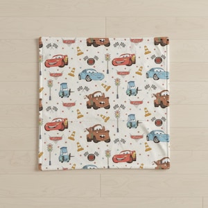 May include: A white blanket featuring a repeating pattern of cartoon cars in red, blue, and brown. The design includes race flags, traffic cones, and traffic lights. The word "Cars" is also printed on the blanket.