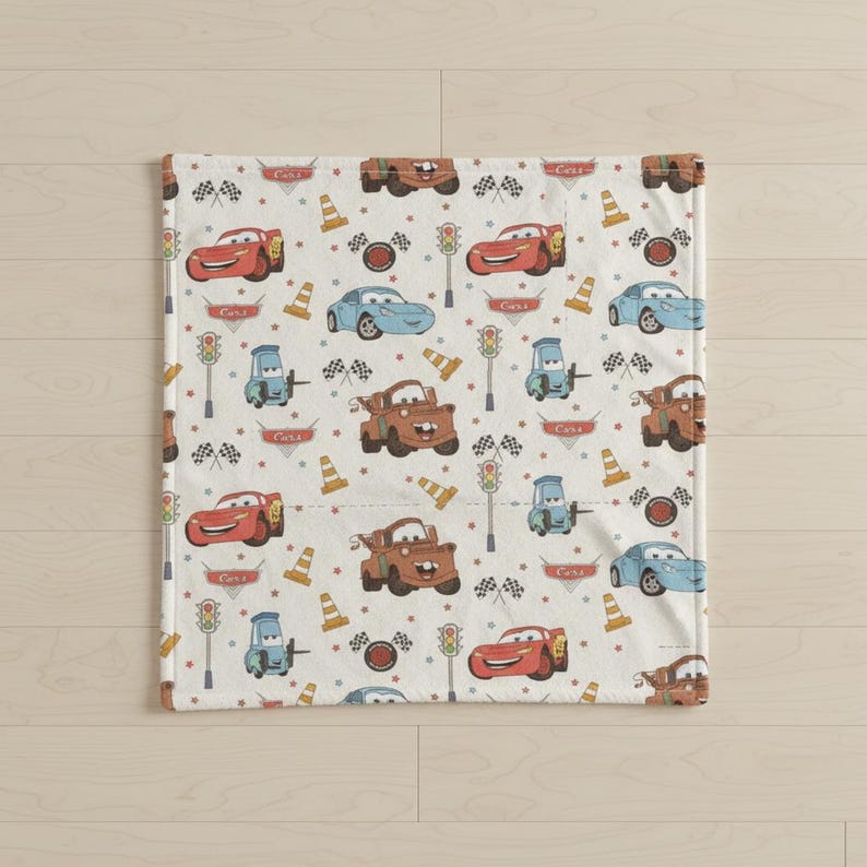 May include: A white blanket featuring a repeating pattern of cartoon cars in red, blue, and brown. The design includes race flags, traffic cones, and traffic lights. The word "Cars" is also printed on the blanket.