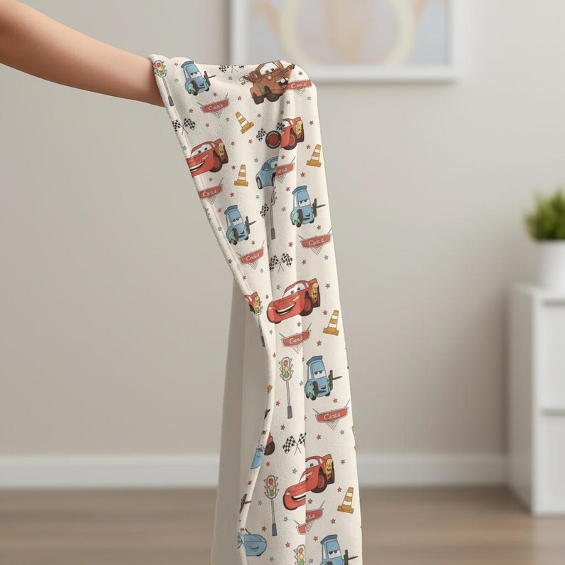 May include: A cream-colored blanket featuring a repeating pattern of cartoon cars, traffic cones, and checkered flags. The cars are in various colors, including red, blue, and brown. The blanket is held up by a person's hand.