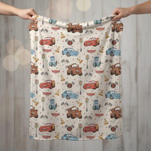 May include: A beige blanket featuring a repeating pattern of cartoon cars in red, blue, and brown. The design includes traffic lights, checkered flags, and traffic cones. The blanket is held up by two hands.