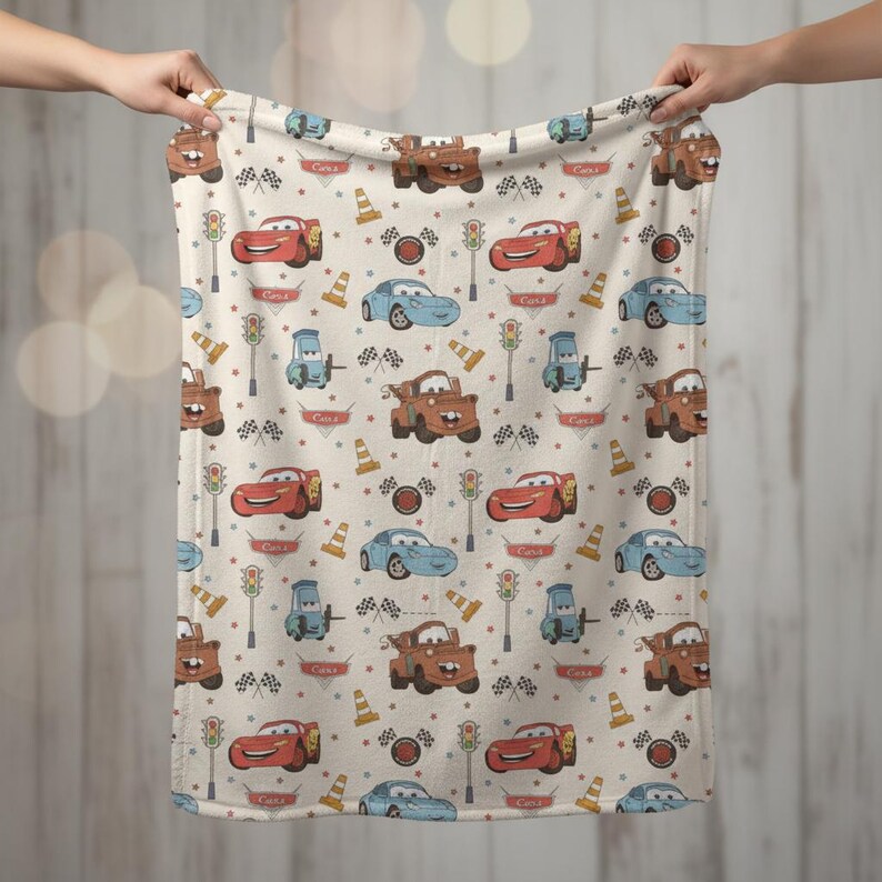 May include: A beige blanket featuring a repeating pattern of cartoon cars in red, blue, and brown. The design includes traffic lights, checkered flags, and traffic cones. The blanket is held up by two hands.