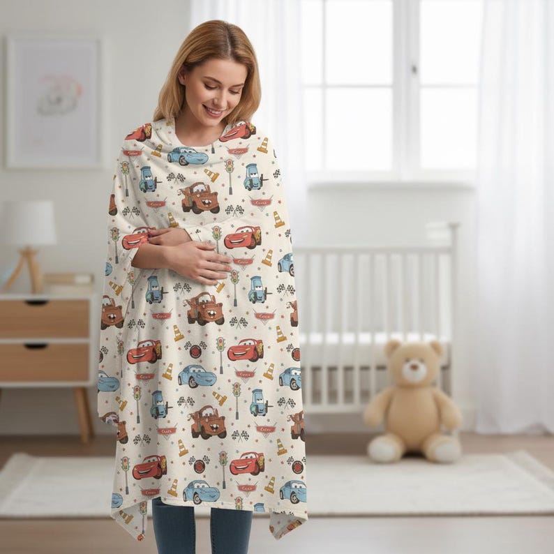May include: A cream-colored baby blanket with a repeating pattern of cartoon cars, traffic lights, and racing flags. The blanket is wrapped around a person. The cars are in shades of red, blue, and brown.
