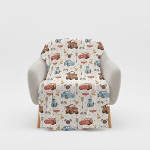 May include: A cream-colored blanket featuring a repeating pattern of cartoon cars, traffic lights, and cones. The cars are in shades of red, blue, and brown. The blanket is draped over a light gray armchair.