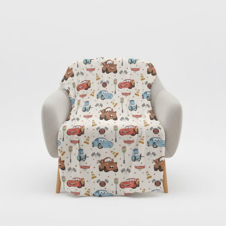 May include: A cream-colored blanket featuring a repeating pattern of cartoon cars, traffic lights, and cones. The cars are in shades of red, blue, and brown. The blanket is draped over a light gray armchair.