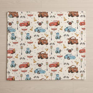 May include: A cream-colored blanket featuring a repeating pattern of cartoon cars in red, blue, and brown. The design includes race flags, traffic lights, and traffic cones. The word "Cars" is printed on some of the cars.