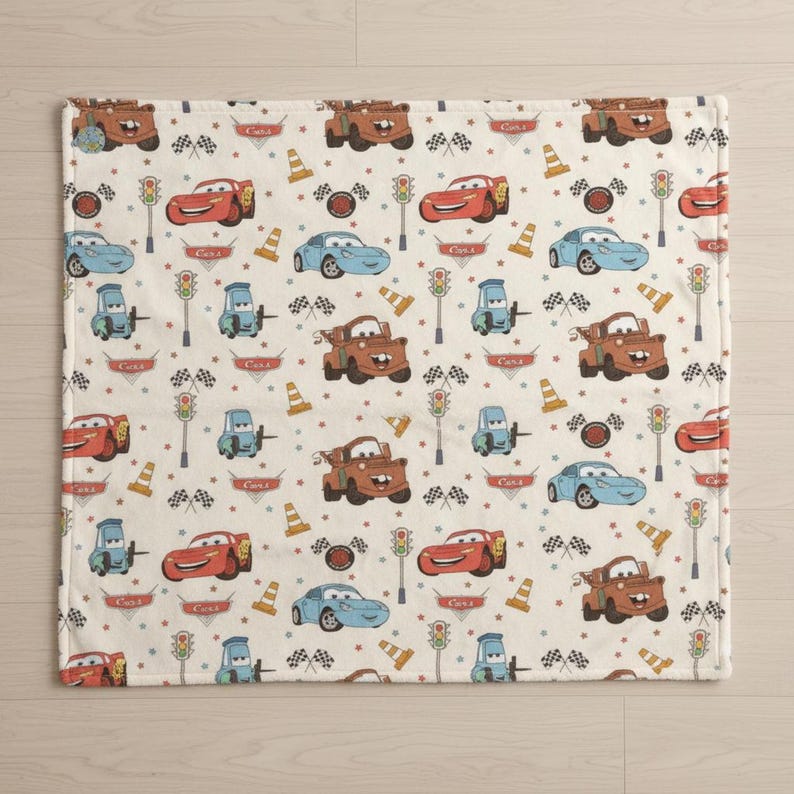 May include: A cream-colored blanket featuring a repeating pattern of cartoon cars in red, blue, and brown. The design includes race flags, traffic lights, and traffic cones. The word "Cars" is printed on some of the cars.