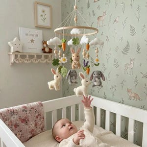 May include: A nursery mobile featuring felted figures of rabbits, white clouds, orange carrots, green clovers, and white flowers suspended from a circular wooden frame. The mobile hangs above a white crib with a baby reaching toward the hanging figures.