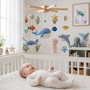 May include: A wooden baby mobile featuring various crocheted sea creatures including a blue whale, a dolphin, a stingray, a jellyfish, a clownfish, a pufferfish, a starfish, and seashells, all suspended from a light-coloured wooden cross frame.