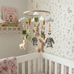 May include: A nursery mobile featuring felted figures of rabbits in white, beige, and gray, suspended from a circular wooden frame. The mobile includes felt clouds, orange carrots, green clover leaves, and white flowers, all hanging from strings with small wooden beads.