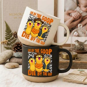 Puede incluir: Dos tazas de cerámica apiladas. La taza superior es de color blanquecino con una base beige, y la taza inferior es negra con una base beige. Cada taza presenta un diseño de cacahuete de dibujos animados y el texto "LIVE BY THE GOOP, DIE BY THE GOOP".