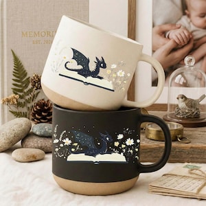 May include: Two ceramic mugs, one cream and one black, stacked. Each mug features a dragon illustration on an open book with floral accents. The mugs have a tan base and a curved handle.