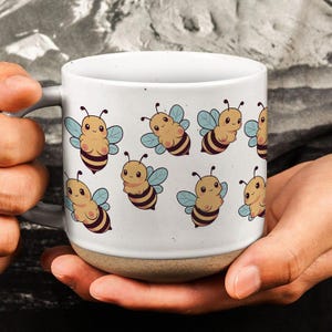 Könnte beinhalten: Eine weiße Keramiktasse mit hellbraunem Boden, die ein sich wiederholendes Muster von Cartoon-Bienen mit hellblauen Flügeln zeigt. Die Tasse wird von den Händen einer Person gehalten.