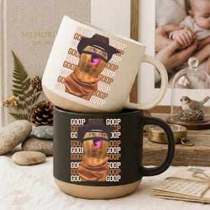 Puede incluir: Dos tazas de cerámica, una crema y otra negra, apiladas. Cada taza presenta un personaje de cacahuete de dibujos animados con sombrero y bufanda, con la palabra "GOOP" repetida verticalmente. Las tazas tienen una base marrón.